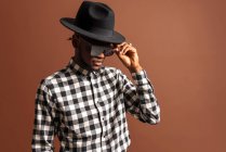 Молодий крутий афроамериканець у картатій сорочці, капелюсі та сонцезахисних окулярах стоячи на коричневому фоні. — стокове фото