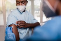 Especialista médico irreconhecível em uniformes de proteção e luvas de látex desinfetando o braço com algodão com álcool para vacinar pacientes afro-americanos do sexo masculino na clínica durante o surto de coronavírus — Fotografia de Stock