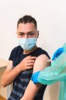 Especialista médica femenina en uniforme protector, guantes de látex y mascarilla facial vacunando a un paciente hispano en clínica durante un brote de coronavirus - foto de stock