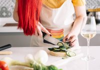 Cultiver homosexuel femelle méconnaissable coupe concombre frais avec couteau tout en préparant des aliments sains à la table de cuisine avec un verre de vin — Photo de stock