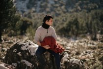 Joven turista masculino en ropa de abrigo contemplando la naturaleza sentado en piedra contra la montaña y mirando hacia otro lado a la luz del sol - foto de stock