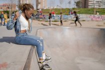Cool fêmea preta com penteado trançado e em patins sentados na rampa no parque de skate e olhando para baixo — Fotografia de Stock