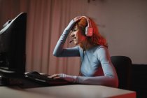 Side view of worried female gamer in headphones playing video game while sitting at table with keyboard — Stock Photo