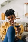 Zufriedene Afroamerikanerin sitzt mit einem Glas Rotwein auf dem Sofa und genießt das Wochenende zu Hause, während sie in die Kamera schaut — Stockfoto