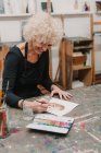 Artista donna in pittura grembiule con acquerelli su carta mentre siede a tavola in laboratorio creativo — Foto stock