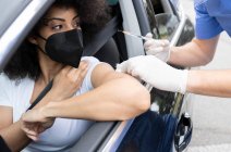 Enfermera irreconocible recortada en guantes de látex y uniforme vacunando a una paciente afroamericana dentro del automóvil en un viaje a través de una clínica móvil durante el brote de coronavirus - foto de stock
