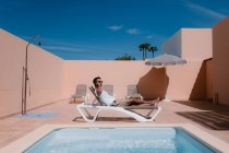 Вид збоку чоловічий фрілансер лежить на шезлонгу біля басейну і виступає на мобільному телефоні під час роботи влітку в сонячний день — стокове фото