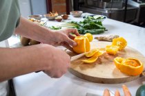 Cultivo irreconocible hembra pelando naranjas jugosas maduras con cuchillo en tabla de cortar de madera en la mesa de la cocina - foto de stock