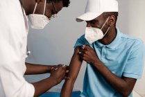 Médico negro lateral en uniforme protector y guantes de látex vacunando a un paciente afroamericano masculino en la clínica durante el brote de coronavirus - foto de stock