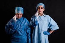 Jóvenes compañeras de trabajo en uniformes médicos quitándose las máscaras faciales mientras están de pie sobre fondo negro en la clínica - foto de stock