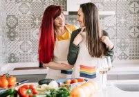 Smiling young woman putting apron on homosexual female beloved at table with assorted fresh vegetables in house — Stock Photo