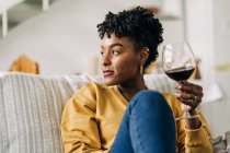 Zufriedene Afroamerikanerin sitzt mit einem Glas Rotwein auf dem Sofa und genießt das Wochenende zu Hause, während sie wegschaut — Stockfoto