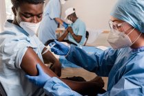Doctora en uniforme protector y guantes de látex vacunando a un paciente afroamericano en la clínica durante el brote de coronavirus - foto de stock