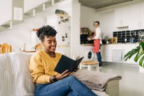 Deliziosa afroamericana lettura femminile interessante libro sul divano sullo sfondo dell'uomo che cucina in cucina — Foto stock