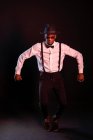 Young masculine ethnic male model in hat and trousers standing dancing on tiptoes while looking at camera on black background — Stock Photo
