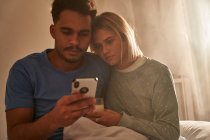 Спокійна багаторасова пара обіймається вранці, сидячи на ліжку і використовуючи смартфон разом — стокове фото