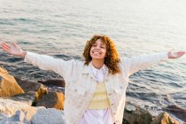 High angle of cheerful African American female standing with outstretched arms on rocks on seashore and enjoying freedom at sunset — Stock Photo