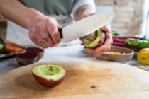 Анонимная женщина с ножом режет спелые половинки авокадо выше доски во время приготовления пищи дома — стоковое фото