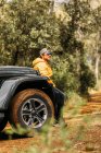 Seitenansicht eines nachdenklichen Abenteurers, der mit Mütze und gelber Jacke vor einem Baumhintergrund vor seinem Geländewagen sitzt — Stockfoto