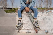 Crop black female with braided hairstyle and in rollerblades sitting on ramp in skate park and looking away — Stock Photo