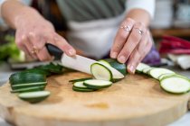 Cultivo de calabacín de corte femenino irreconocible con cuchillo mientras se prepara el almuerzo en la mesa de la cocina en casa - foto de stock
