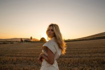 Мирная женщина в элегантном платье стоит на сухом поле в сельской местности и смотрит в камеру — стоковое фото