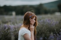 Vista lateral de hembra suave con flores en el pelo sentado en el campo de lavanda floreciente y disfrutando de la naturaleza con los ojos cerrados - foto de stock