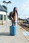 Vista lateral do viajante asiático feminino com mala em pé na plataforma da estação ferroviária enquanto espera o trem — Fotografia de Stock
