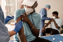 Gestutzter Arzt in Schutzuniform und Latexhandschuhen impft während des Coronavirus-Ausbruchs unkenntlich gewordenen afroamerikanischen Patienten in Klinik — Stockfoto