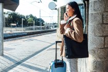 Боковой вид контента: женщина стоит с чемоданом на платформе на вокзале и разговаривает по смартфону — стоковое фото