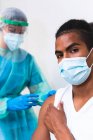 Especialista médica femenina en uniforme protector, guantes de látex y mascarilla facial vacunando a un paciente afroamericano en clínica durante el brote de coronavirus - foto de stock