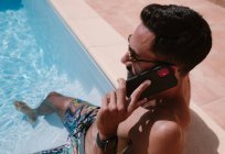 С вышестоящего серьезного безрубашечного мужчины-фрилансера, сидящего у бассейна с ногами в воде и разговаривающего по мобильному телефону во время удаленной работы летом — стоковое фото