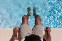 Вид сверху неузнаваемого мужчины-фрилансера, сидящего у бассейна с ногами в воде во время телеработы летом — стоковое фото