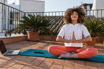 Tranquila mujer afroamericana sentada en Padmasana con gesto Namaste y meditando con los ojos cerrados mientras practica yoga en la azotea - foto de stock