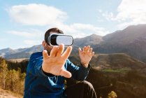Curieux voyageur masculin assis sur la colline et l'expérience de la réalité virtuelle dans les lunettes de soleil dans les montagnes le jour ensoleillé — Photo de stock