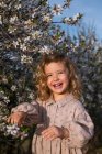 Adorable niño sonriente en vestido de pie cerca del árbol en flor con flores en el parque de primavera y mirando hacia otro lado - foto de stock