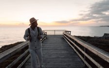 Hombre con mochila y sombrero de pie en una pasarela mirando al mar - foto de stock