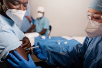 Ärztin in Schutzuniform und Latexhandschuhen impft während des Coronavirus-Ausbruchs einen afroamerikanischen Patienten in der Klinik — Stockfoto