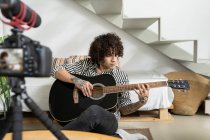 Молодий татуйований гітарист грає на акустичній гітарі під час запису відео на фотокамеру в домашній кімнаті — стокове фото