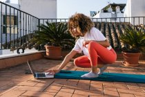 Сфокусована афроамериканська жінка з кучерявим волоссям вибирає онлайн-посібник на ноутбуці, сидячи на маті на даху і готуючись до уроку йоги. — стокове фото