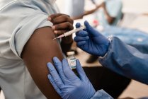 Обрезанный врач в защитной форме и латексных перчатках вакцинирует неузнаваемого пациента-афроамериканца в клинике во время вспышки коронавируса — стоковое фото