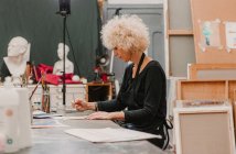 Artista femenina enfocada sentada a la mesa y pintando con acuarelas sobre papel mientras trabaja en un taller creativo - foto de stock