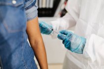 Especialista médico masculino irreconocible en uniforme protector y guantes de látex vacunando a una paciente afroamericana en la clínica durante el brote de coronavirus - foto de stock