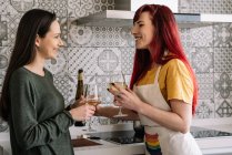 Seitenansicht junger, lächelnder homosexueller Frauen mit Gläsern und einer Flasche Weißwein, die im Haus sprechen, während sie einander anschauen — Stockfoto