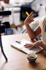 Zugeschnittene unkenntliche Vloggerin mit Notizbuch sitzt am Tisch mit Fotokamera auf Stativ in der Küche — Stockfoto