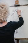Обратный вид анонимной художницы, создающей рисунок человека карандашом, стоя у мольберта в студии — стоковое фото