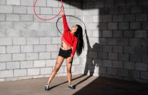 Joven mujer tatuada en ropa deportiva girando hula hoop mientras baila contra paredes de ladrillo con sombras y mirando hacia adelante a la luz del sol - foto de stock