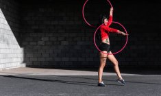Giovane donna tatuata in activewear vorticoso hula hoop mentre balla contro muri di mattoni con ombre e guardando avanti alla luce del sole — Foto stock