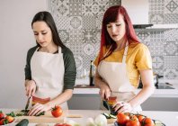 Homosexual girlfriends cutting cucumber while preparing healthy vegetarian food at table in house — Stock Photo