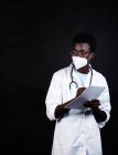 Médico afroamericano en máscara protectora y uniforme blanco tomando notas en el portapapeles mientras mira hacia otro lado sobre fondo negro - foto de stock